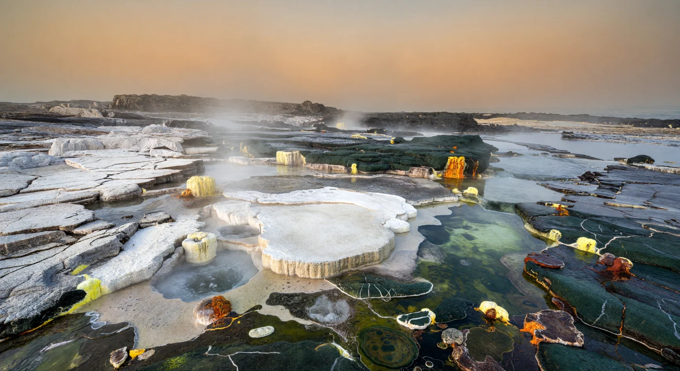 約29億〜27億年前の太古代後期、安定しはじめたクラトン縁辺の熱水地帯では、TTG質花崗質岩や暗色のグリーンストーンが割れた岩棚をなし、白いシリカ焼結堆積物、赤い鉄酸化物、黄色い硫黄が噴気孔のまわりを鮮やかに縁どっていました。湯気の立ちこめる湿った岩肌にだけ、緑黒色や褐紫色のごく薄い微生物マットが張りつき、乾いた地表は植物も動物もいない完全な裸岩の世界です。こうした微生物群はシアノバクテリアやアナオキシック光合成細菌、化学合成細菌を含んでいた可能性があり、海へ流れこむ熱水は初期大陸の風化と生命の営みを結びつける、地球史でもきわめて古い陸上環境を物語っています。